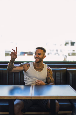 A vertical shot of a young smiling caucasian guy with cool tattoos sitting in the cafe and calling a waiterの写真素材