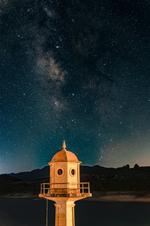 A vertical shot of a lighthouse with the silhouettes of mountains under a dark blue starry skyの写真素材