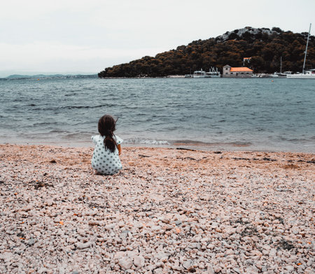 A vertical shot of a little girl sitting alone on the beach looking at the blue seaの写真素材
