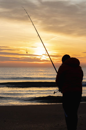A vertical shot of a fisherman with a fishing pole on the shore on a golden sunsetの写真素材