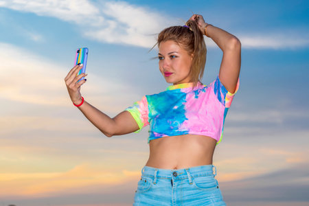 Young woman in bright tracksuit stands against background of an evening sunset and takes pictures of herself on mobile phone. Outdoor recreation.の写真素材