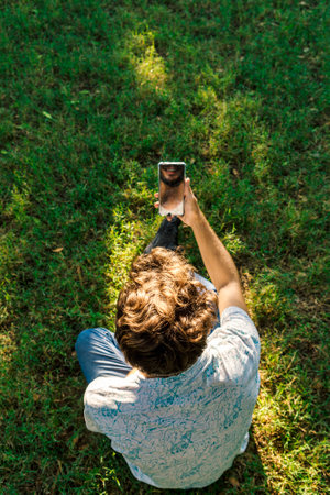 A vertical shot of a young Spanish guy from above, sitting on the grass in a park and taking a selfieの写真素材