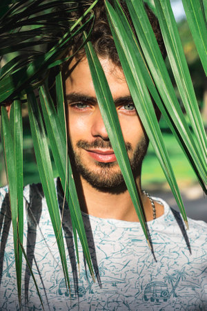 A vertical shot of a handsome Spanish guy looking at the camera with a smile from behind long tropical leavesの写真素材