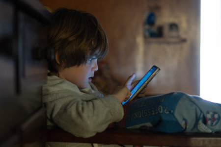 A closeup of a little boy looking at the smartphone in the bed.