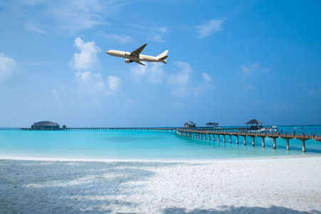 A shot of a white airplane flying over a tropical sea with a sandy beach on a sunny dayの写真素材