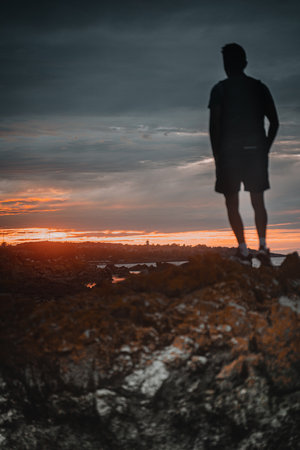 A vertical shot of the silhouette of a male standing on the rock looking at sunsetの写真素材