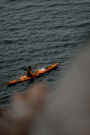 A vertical shot of a person with an orange kayak in the gray wavy sea with a blurred rock in the...