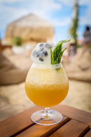 A closeup of a delicious orange cocktail with iced blueberry and mint on a sunny summer beach