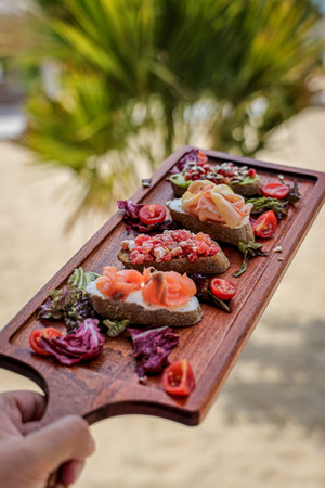 A vertical shot of delicious appetizers on a wooden tray at the beach on a sunny dayの写真素材