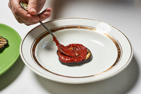 A hand adding tomato sauce on eggplant with spoonの写真素材