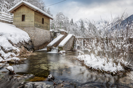 A beautiful shot of a house in the middle of a forest near a river covered in snow