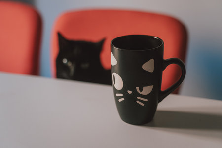A black mug with a cat face on the table with a black cat near itの写真素材