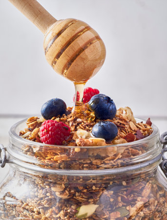 A vertical shot of honey pouring on an oatmeal jar with berries against a white blurry backgroundの写真素材