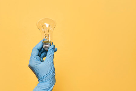 A person's hand with a medical glove holding a light bulb on a yellow background with copy spacの写真素材