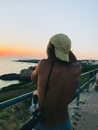 A shirtless young man taking a picture of the sea during the sunset with a camera