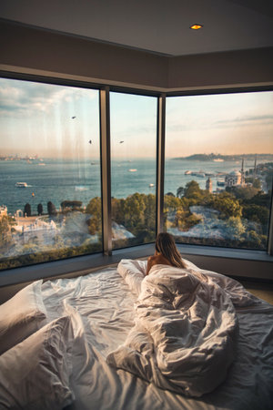 A rear view of a female lying on bed looking at the beach through the glass windows