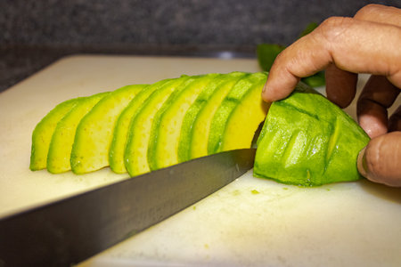 A male's hand slicing an avocado with a knife on a white cutting board