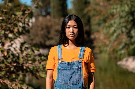 A pretty East Asian lady with dark hair and jeans overalls posing in the park
