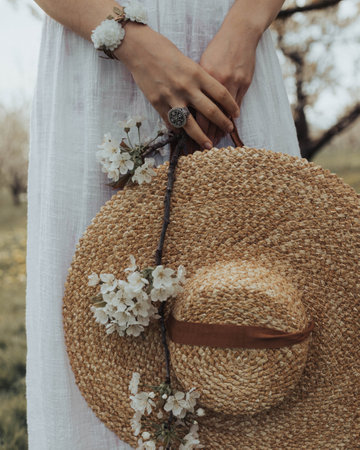 A vertical shot of a woman holding a wicker hat and a branch with flowersの写真素材