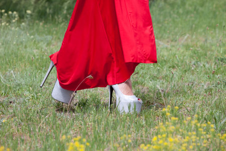 A closeup shot of a person wearing a red long dress and white high heels walking in a parkの写真素材