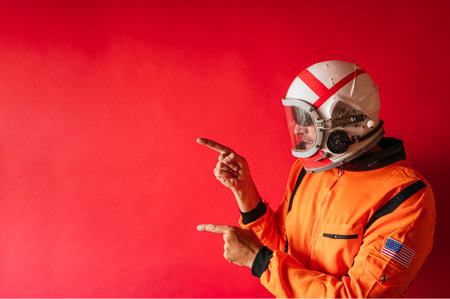 An astronaut with a helmet and an orange space suit pointing on a red background with copy space