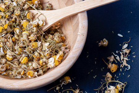 A closeup shot of dry chamomile petals in a wooden pot with wooden spoon on a black fabricの写真素材