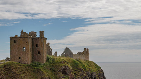 The famous historic ruined medieval fortress of Dunnottar Castle on a rocky headland under a blue cloudy skyの写真素材