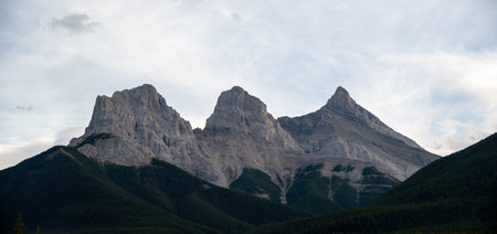 A mesmerizing view of three tops of mountains in The Three Sisters, Alberta, Canada under a cloudy skyの写真素材