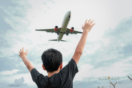 A shot of a little boy with his hands raised under a flying airplane with a cloudy sky backgroundの写真素材