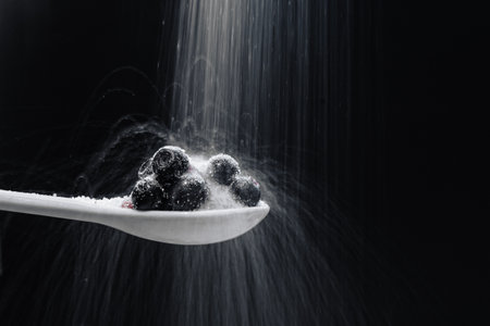 A closeup shot of pouring white sugar on blueberries in a white spoon against a black background