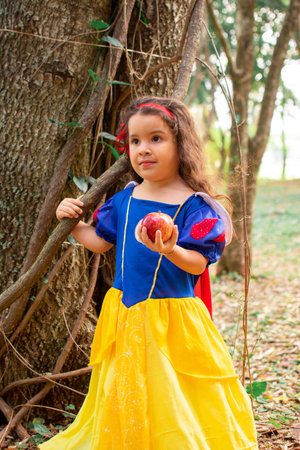 A cute little Hispanic girl wearing a Snow White dress and holding an apple in a forestの写真素材