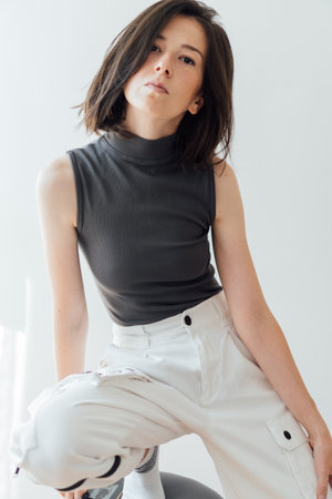 A young Romanian brunette woman wearing a sleeveless jumper and white pants, posing in a bright roomの写真素材
