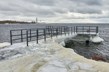 The pier is covered with a thick layer of ice. A lighthouse is visible in the distance. Cloudy weather.の写真素材