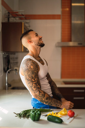 A side portrait of a young tattoed guy standing in the kitchen beside the vegetablesの写真素材