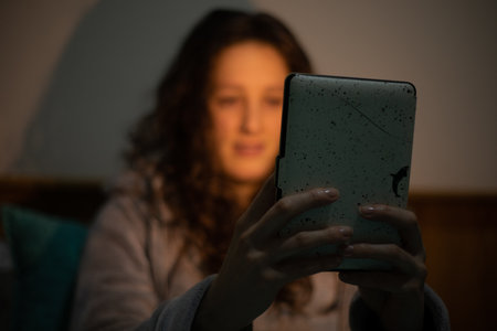 A closeup of a young woman in a hoodie sitting in the darkness and looking at the tablet.の写真素材