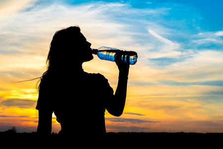 Silhouette of a young girl on the background of a summer sunset. Drinks water from a plastic bottle.の写真素材