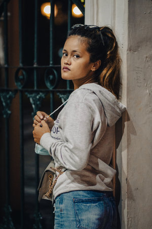 A vertical shot of a young Southeast Asian girl on the street leaning against a wall at night in...