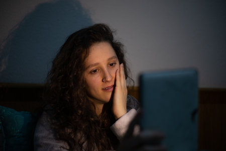 A closeup of a young woman in a hoodie sitting in the darkness and looking at the tablet.の写真素材