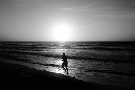 A grayscale of a silhouette of a man running across the beach on a sunriseの写真素材