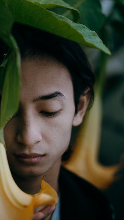 A vertical shot of a young handsome Indonesian man posing in nature with leaves and flowers