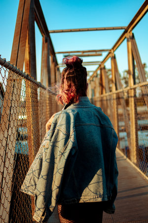 A vertical shot of a cool young lady with pink hair and gothic style clothing on a bridge