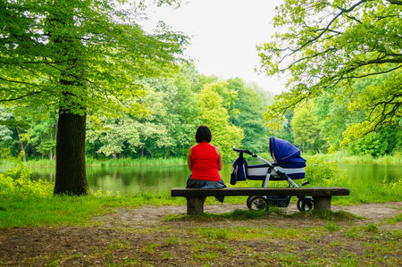 A  Caucasian woman sitting on a wooden bench next to a baby buggy watching a lake in the gardenの写真素材