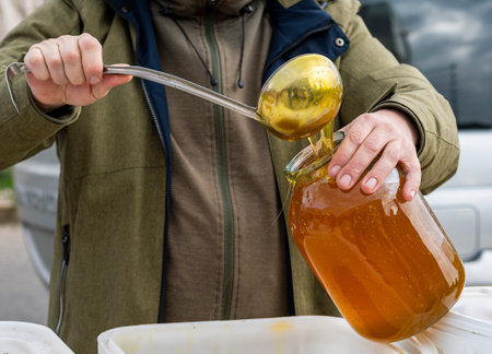 The man's cut-off hands are pouring a mass of bee honey into a glass jar using a ladle. Agricultural Products Fair.の写真素材