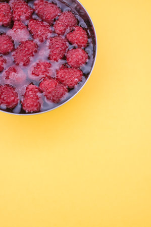 A vertical shot of a bowl with water and raspberries in it on the yellow background
