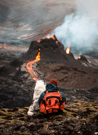 A vertical shot of a person with a dog looking at an active volcano with flowing lavaの写真素材