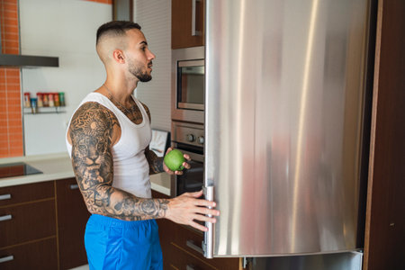 A horizontal shot of a young tattooed guy taking an apple out of the fridgeの写真素材
