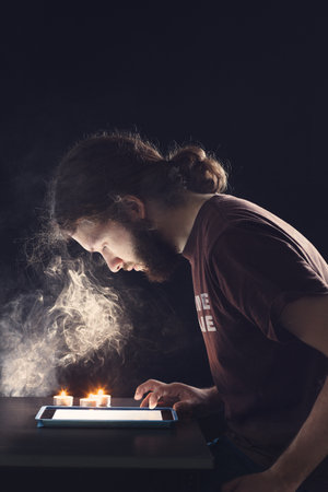 A portrait of an adult man with a beard and long hair using his tablet on the table with candles on...
