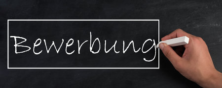 The word application is standing in german language on a chalkboard, hand with white chalkの写真素材