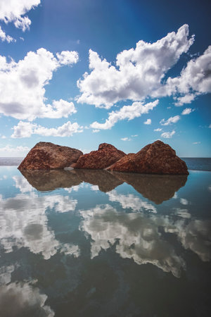 A vertical shot of three rocks in the sea under a blue cloudy sky and sunlightの写真素材