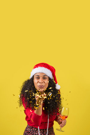 A young Hispanic female wearing a Santa hat, blowing on shiny golden paper while holding a wineglassの写真素材